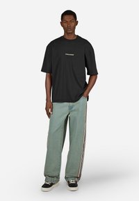 Młody mężczyzna ubrany w czarną koszulkę "PEGADOR", jasnoniebieskie szerokie dżinsy z bocznymi paskami i czarne trampki z białymi podeszwami i sznurówkami.