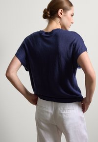 Marineblaues Leinen-T-Shirt mit kurzen Ärmeln und lockerer Passform. Der Stoff hat eine leichte Textur. Kombiniert mit hellen Leinenhosen, von hinten gezeigt.