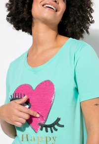 Mintgrøn T-shirt med korte ærmer, der har et stort pink glitrende hjerte med øjenvipper og ordet "Glad" i guld nedenfor. Blød stofstruktur.