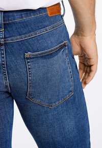 Primo piano di una persona che indossa jeans di denim blu con una tasca posteriore visibile e una patch del marchio in pelle, mano appoggiata vicino all'anca.