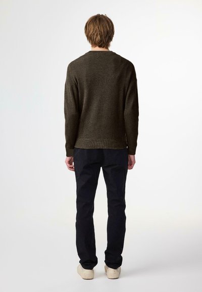 Skarpetkowy sweter w oliwkowym kolorze z długimi rękawami, okrągłym dekoltem i prążkowanymi mankietami, w zestawieniu z czarnymi spodniami i jasnymi butami.