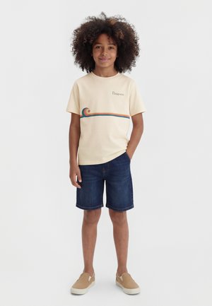 Παιδί με σγουρά μαλλιά στέκεται με το ένα χέρι στην τσέπη, φορώντας μπεζ μπλούζα Patagonia με σχέδιο κύματος, σορτς από τζιν και καφέ παπούτσια τύπου slip-on.