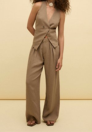 Femme debout portant un gilet sans manches à fines rayures marron, un pantalon ample assorti, un collier ras du cou noir avec une fleur blanche, et des sandales marron.