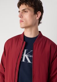 Maroon bomber jacket with a smooth texture, featuring a standing collar and zip closure, layered over a navy sweatshirt with a logo print.