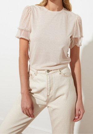 Vrouw draagt een lichtbeige blouse met doorschijnende pofmouwen en hooggetailleerde off-white jeans tegen een effen achtergrond.