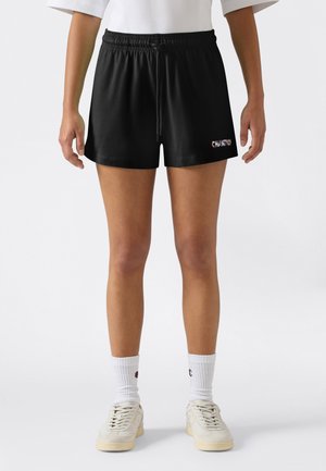 Vrouw draagt zwarte shorts met elastische taille en trekkoord, witte kousen en witte sneakers, staat met armen ontspannen langs het lichaam.