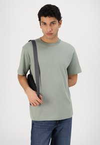Short-sleeve, sage green T-shirt in cotton blend. Features a crew neckline and relaxed fit. Paired with dark jeans and a black crossbody bag.