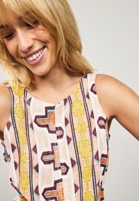 Haut sans manches à motifs combinant des dessins géométriques jaunes, oranges et bruns avec des accents bleus sur fond blanc. Texture douce.