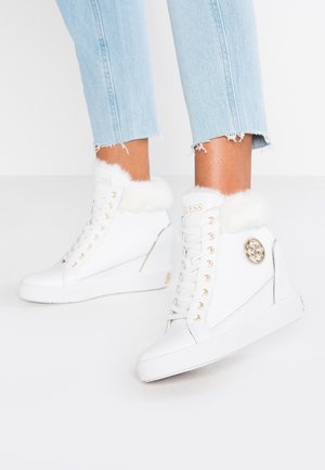 Białe sneakersy na wysokiej koturnie z futrzanym obszyciem, złotymi przelotkami i emblematem logo, noszone z postrzępionymi jasnoniebieskimi dżinsami.