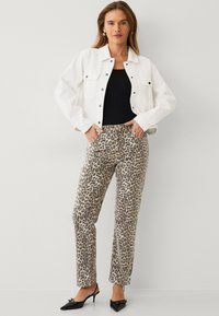 Hvid denimjakke med trykknapper, sort tanktop og beige leopardmønstrede bukser. Sorte hæle fuldender outfittet. Simpelt baggrund.