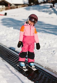 Dítě stojí na lyžařském dopravníku v růžovém a oranžovém sněhovém obleku s černými rukavicemi, na hlavě má helmu a brýle, obklopeno sněhem.