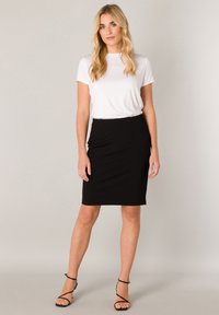 Schwarzer Bleistiftrock aus elastischem Stoff, kombiniert mit einem weißen T-Shirt mit Rundhalsausschnitt; das Model trägt schwarze Riemchenpumps und steht vor einem neutralen Hintergrund.