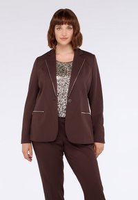 Hnědý blazer se strukturovaným designem, který má kontrastní stříbrné zipové detaily a zavírání na předním tlačítku, nošený přes top se sequiny.