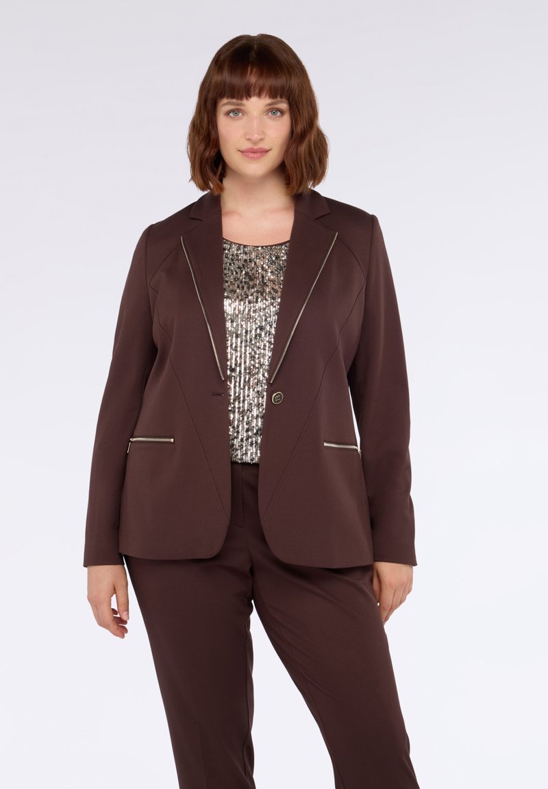 Hnědý blazer se strukturovaným designem, který má kontrastní stříbrné zipové detaily a zavírání na předním tlačítku, nošený přes top se sequiny.