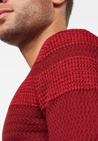Maglione rosso lavorato a maglia con motivi texturizzati e collo a coste, che mostra un primo piano della spalla e della parte superiore del torace.