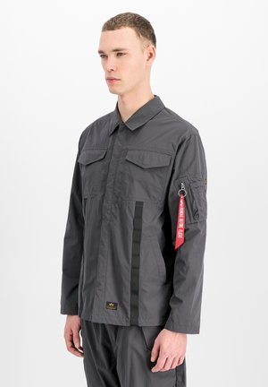 Mann trägt graue Jacke mit Klappentaschen an der Brust und rotem "Remove Before Flight" Anhänger an der Reißverschlusstasche am linken Ärmel.