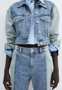 Veste en denim aux teintes bleu clair et bleu moyen, au design cropped, avec deux poches poitrine et des accessoires en argent. Associée à un jean taille haute bleu clair et à une chemise rayée.