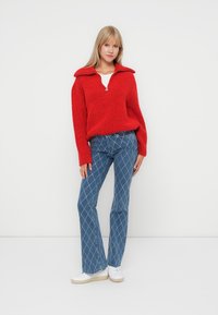 Rode, gestructureerde pullover met een kraag en rits; gecombineerd met blauwe, gedessineerde flare jeans met diamantpatronen; witte sneakers.