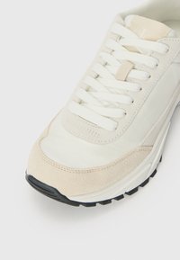Light beige sneaker with a mix of leather and suede materials, featuring white laces, a cushioned sole, and a black rubber outsole.