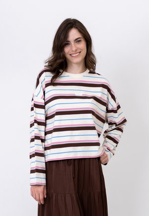 Donna con lunghi capelli castani che indossa una camicia bianca a maniche lunghe con strisce orizzontali marroni, rosa e blu e una gonna marrone, sorridente.
