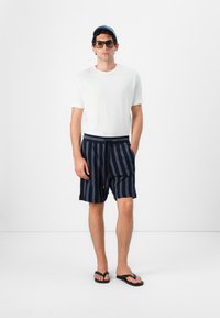 Biele tričko, námornícky pruhované šortky a čierne žabky. Model má na sebe veľké slnečné okuliare a modrú čiapku. Jednoduchý, neformálny letný outfit.