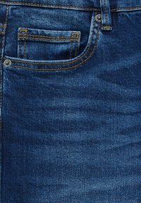 Närbild av en blå denimbyxas ficka med orange sömmar, liten myntficka och metallnitar.