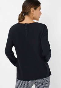 Zwarte langemouwen top met een ronde halslijn en een ritsdetail aan de achterkant. Soepele textuur, relaxte pasvorm, gecombineerd met gemusterde broeken.