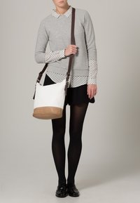 Borsa a spalla in tela bianca con una base marrone, dotata di una lunga tracolla marrone. Il modello indossa un maglione grigio sopra una camicia a pois.