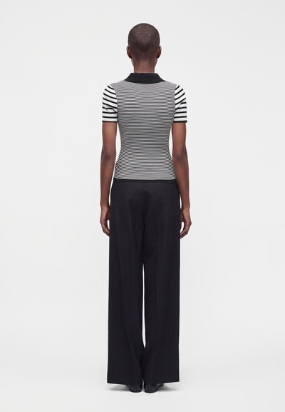 Personne aux cheveux courts portant un haut à manches courtes rayé noir et blanc et un pantalon noir à jambes larges, debout de dos sur un fond uni.