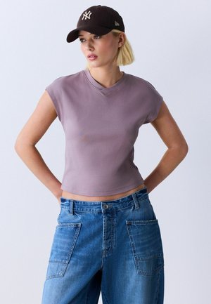 Donna che indossa un cappellino nero dei New York Yankees, un top senza maniche color malva e jeans in denim blu a vita alta con grandi tasche frontali, in piedi con le mani dietro la schiena.