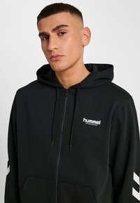 Zwarte rits hoodie van een katoenmix. Beschikt over een voorzak, een capuchon met aantrekkoord en een wit logo op de borst en mouw.