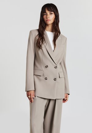 Jeune femme aux longs cheveux bruns portant un blazer beige à double boutonnage et un pantalon large assorti, se tenant devant un fond clair uni.
