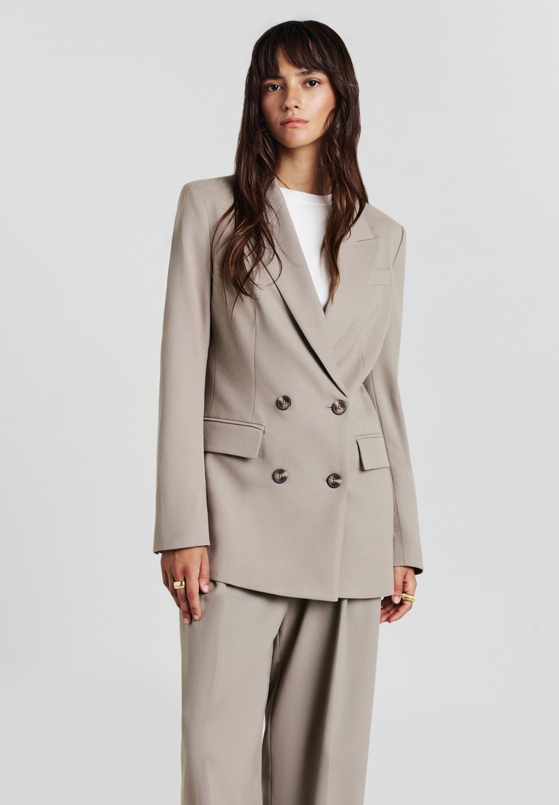 Giovane donna con lunghi capelli castani indossa un blazer beige doppiopetto e pantaloni larghi abbinati, in piedi di fronte a uno sfondo chiaro uniforme.