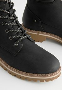 Botas negras con cordones, hechas de cuero sintético texturizado. Cuentan con cordones moteados en contraste y una suela gruesa y resistente con un patrón de banda de rodadura.