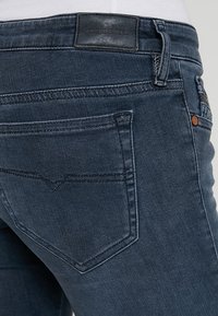 Mörka denimjeans med en slät textur, med en läderlogga vid midjan, två bakfickor och kontrasterande sömmar.