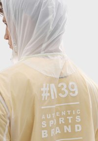 Veste imperméable translucide jaune clair avec une capuche en mesh, comportant un texte blanc : "#39 MARQUE SPORTIVE AUTHENTIQUE" au dos.