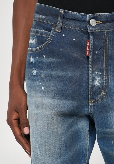 Jeans de denim azul con detalles desgastados, áreas descoloridas y una etiqueta bordada en rojo. Remaches de cobre y diseño clásico de cinco bolsillos.