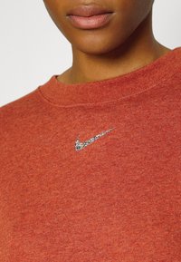 Primo piano di una persona che indossa una maglietta a girocollo rossa con un piccolo logo Nike a forma di baffo in argento sul petto.