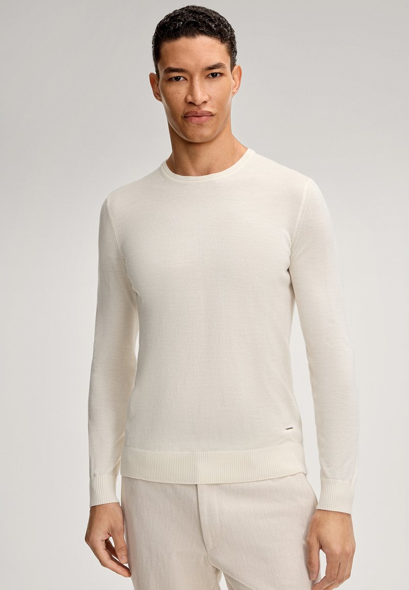 Maglione beige chiaro a maniche lunghe con scollo rotondo, realizzato in tessuto morbido. Presenta polsini e orlo a coste, con un dettaglio di branding minimo.