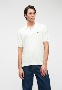 Polo shirt blanc à manches courtes avec un tissu texturé, patte de boutonnage à deux boutons et un petit logo vert sur la poitrine, associé à un jean en denim foncé.