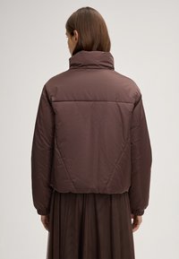 Giacca a vento marrone con colletto alto, polsini elastici e design cropped. Il materiale appare liscio e lucido.