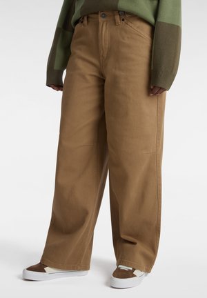 Osoba ubrana w szerokie brązowe spodnie, zielono-brązowy sweter z długim rękawem oraz białe trampki z brązowymi i beżowymi akcentami.