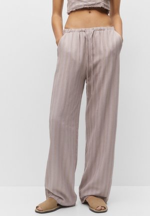 Persoon die losse, licht mauve gestreepte broek en sandalen draagt, met handen in de zakken en een cropped top die de buik laat zien.