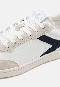 Weißer Sneaker mit beigen Wildlederelementen und navyblauen Details. Verfügt über flache Schnürsenkel, eine strukturierte Sohle und einen eingeprägten Markennamen.