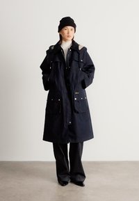 Μακρύ παρκά σε ναυτικό μπλε χρώμα με κλείσιμο με φερμουάρ και κουμπιά, δύο μπροστινές τσέπες και κουκούλα. Κατασκευασμένο από ανθεκτικό ύφασμα, με ασημένιες λεπτομέρειες.