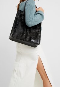 Sac cabas noir en similicuir à texture crocodile, avec une forme rectangulaire et des anses solides, tenu devant un fond clair.