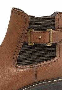 Brown leather ankle boot with a textured brown elastic panel, accented by a metallic buckle at the side and contrasting stitching details.
