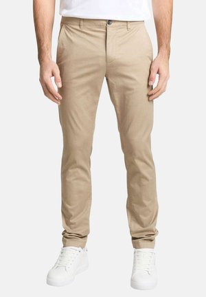 Man wearing slim-fit beige chinos and white sneakers standing against a plain background.