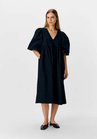 Robe midi noire en coton avec un col en V, des manches bouffantes et une taille froncée ; associée  à des chaussures plates bordeaux. Texture lisse, design simple.