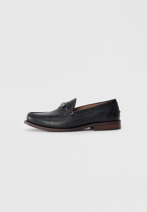 Mocasines de cuero negro con punta redondeada, hardware decorativo en la parte delantera y una suela marrón contrastante. Textura suave y diseño fácil de poner.